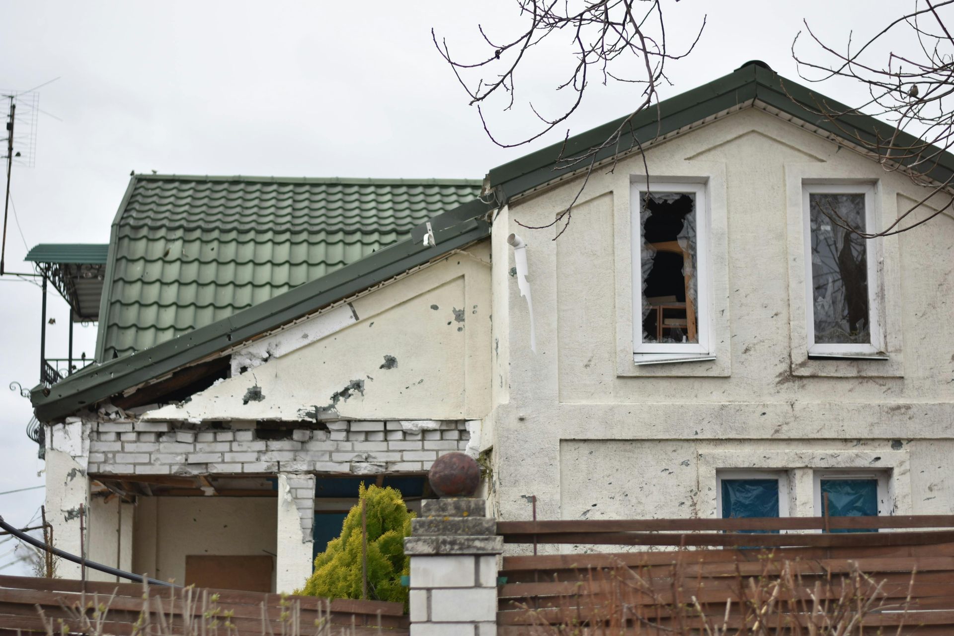 Damaged house with broken windows and a green tiled roof behind a fence