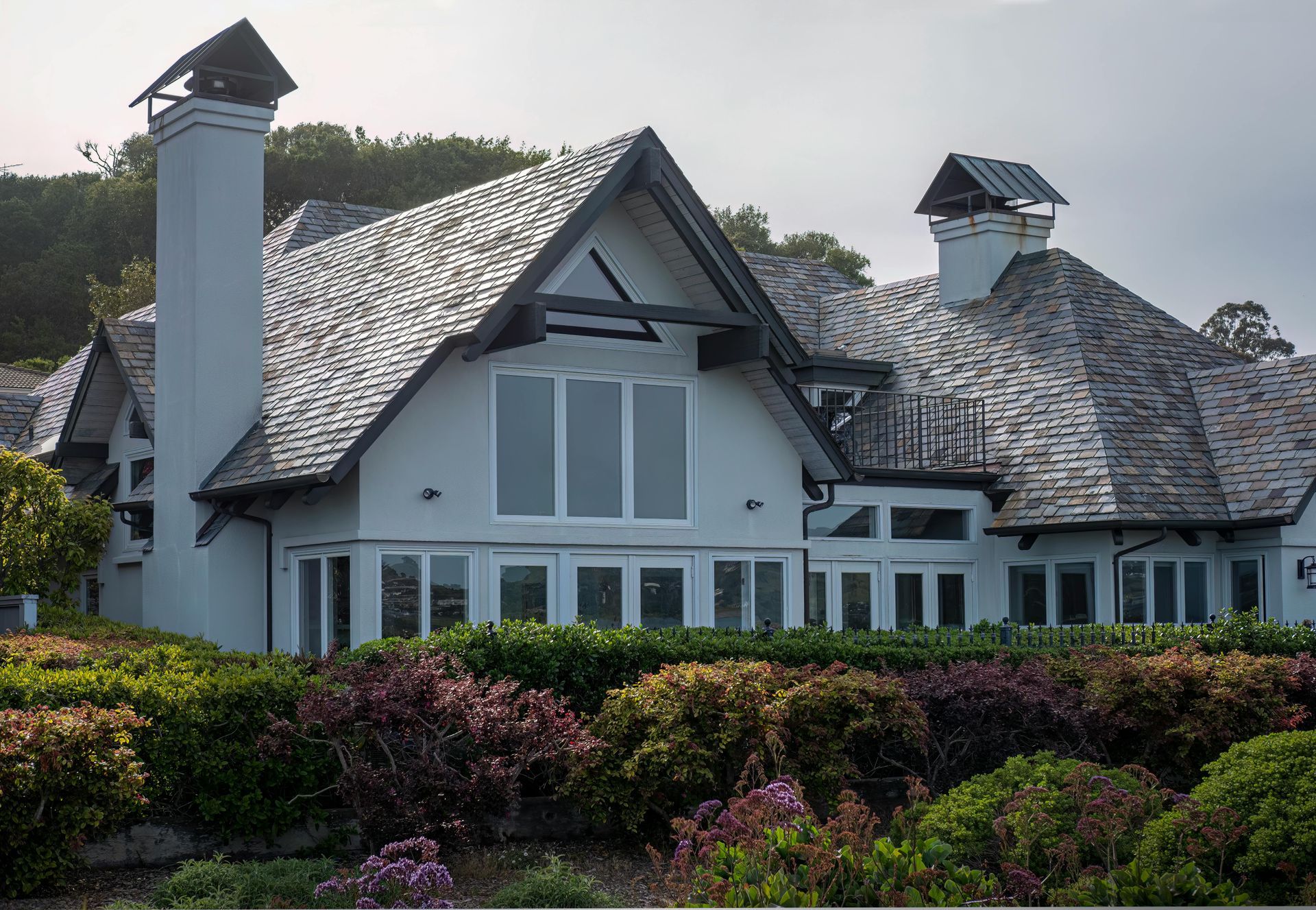 Large gray house with steep roof and dormers, surrounded by lush green and purple shrubs