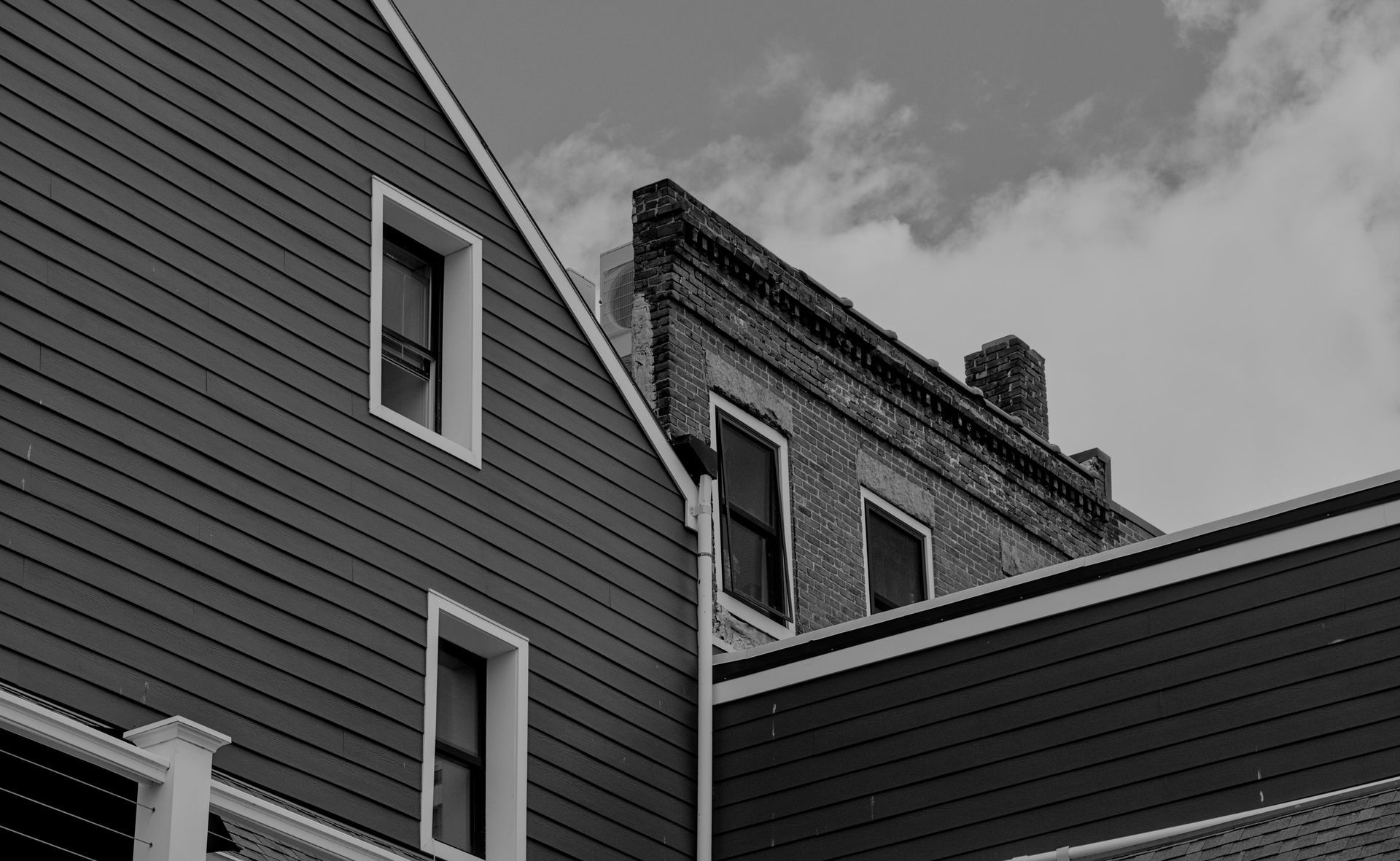 Black-and-white view of adjacent house rooftops and siding against a cloudy sky