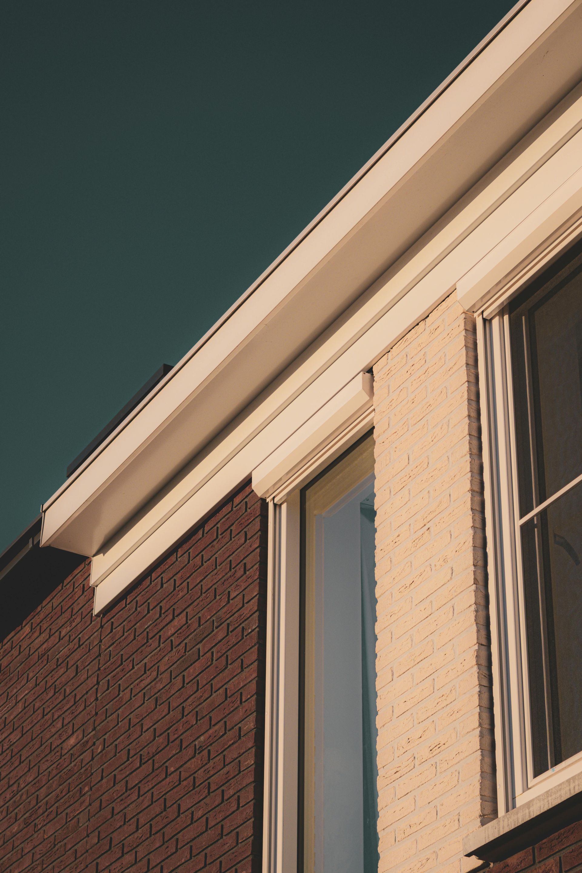 Close-up of a brick building corner with cream trim and a window against a teal sky