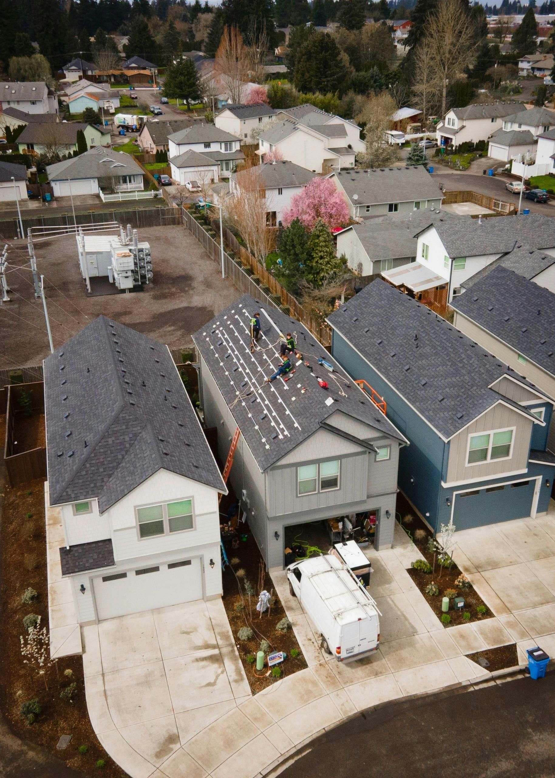 Aerial view of suburban houses with gray roofs on a curved street, one with pink flowering tree in front.