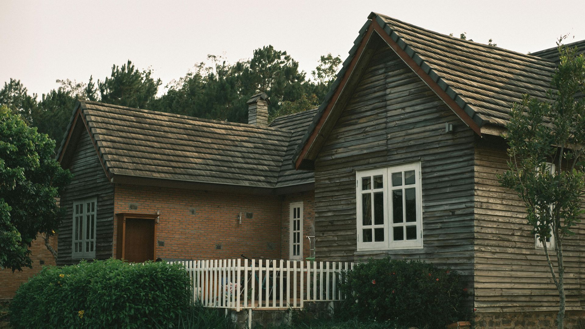 Old rustic cottage with weathered wood siding, white-framed windows, and a white picket fence amid trees