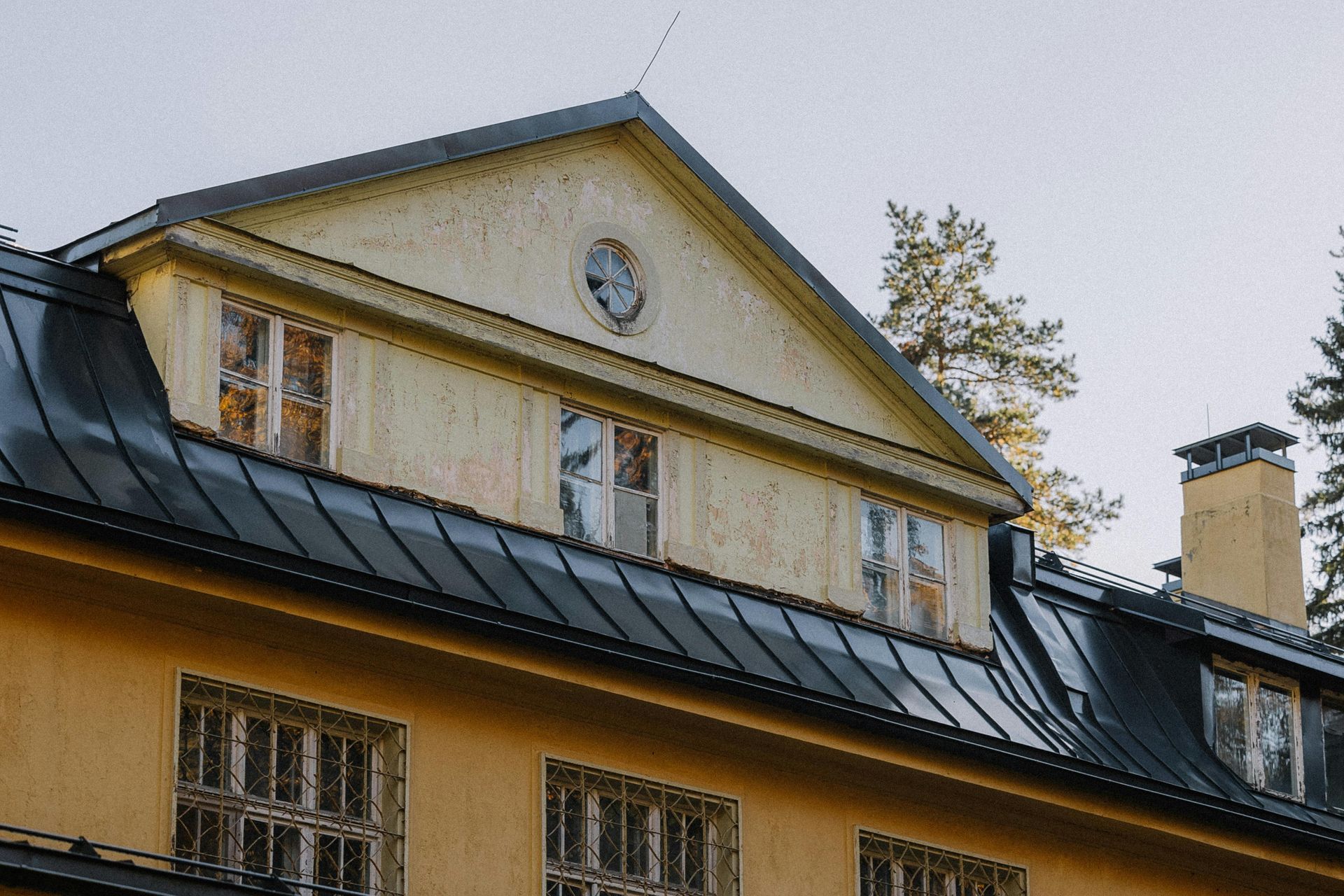 Yellow building facade with a dark metal roof, tall windows, and a clock under the gable roofline
