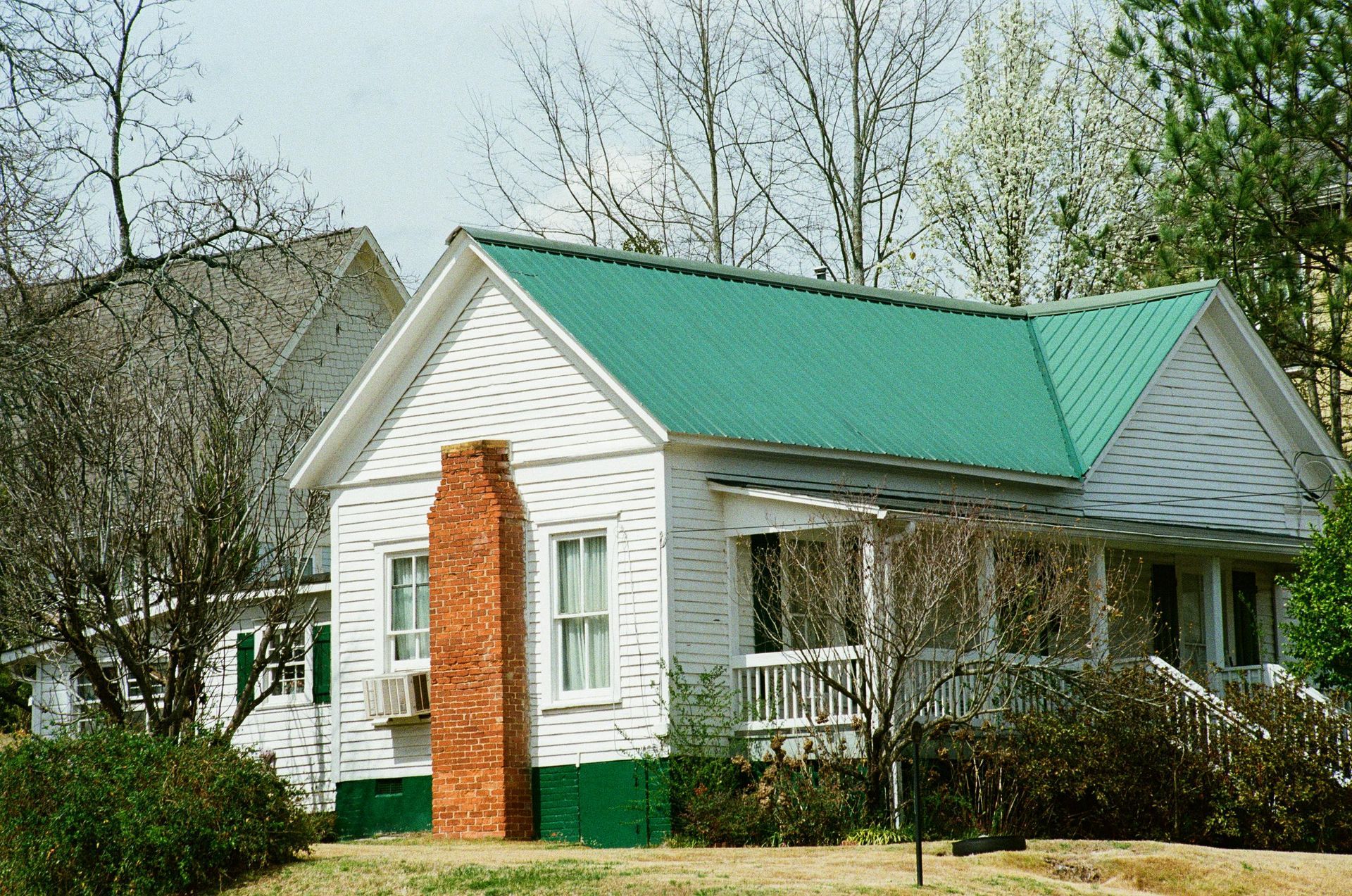 Small white house with a green roof and brick chimney, surrounded by trees and bushes.