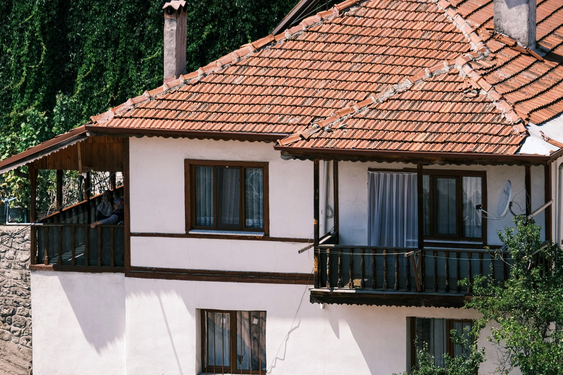 Two-story house with red tiled roof, white walls, and dark wooden balconies amid green trees