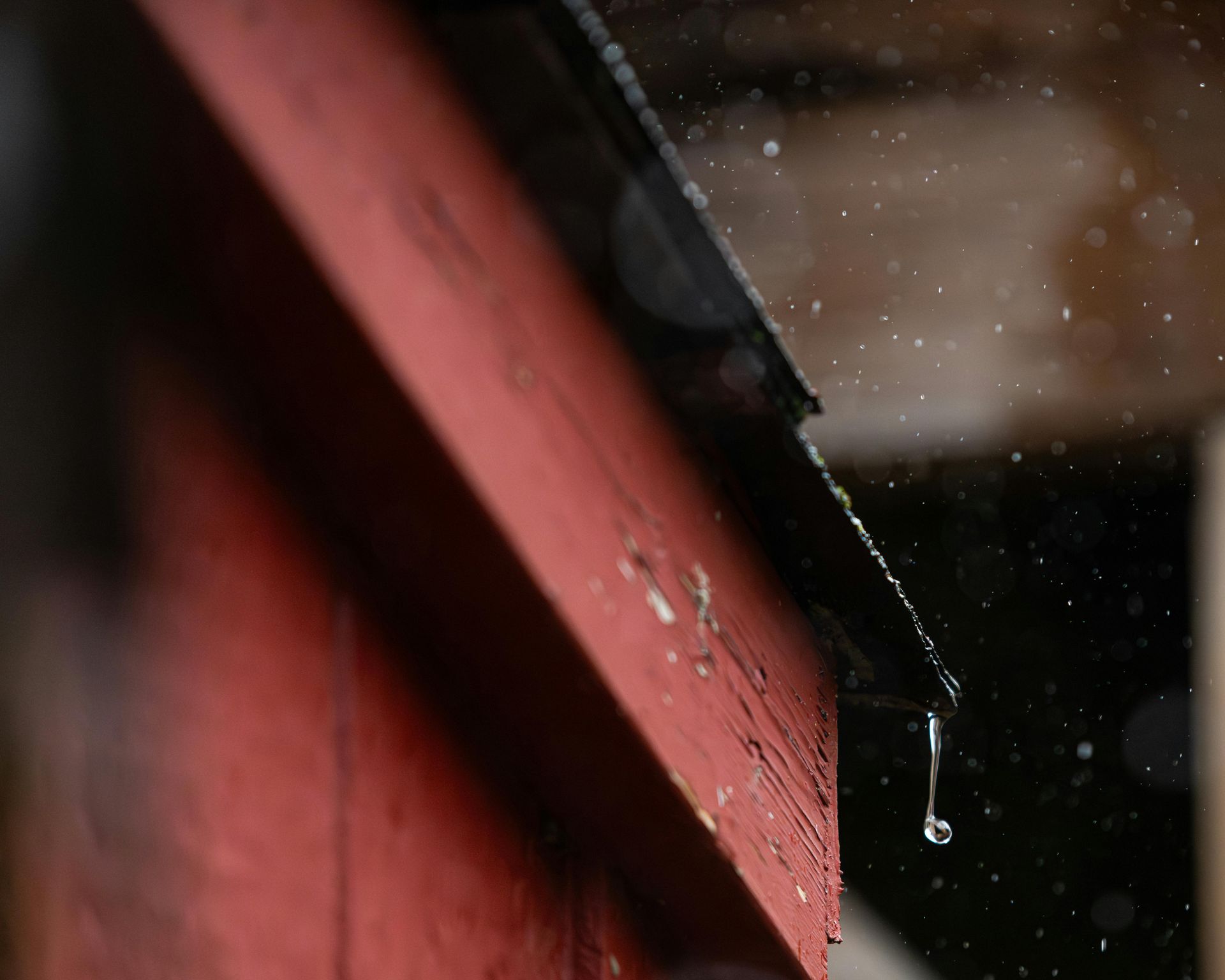 Close-up of a red wooden roof edge with rainwater dripping against a blurred background