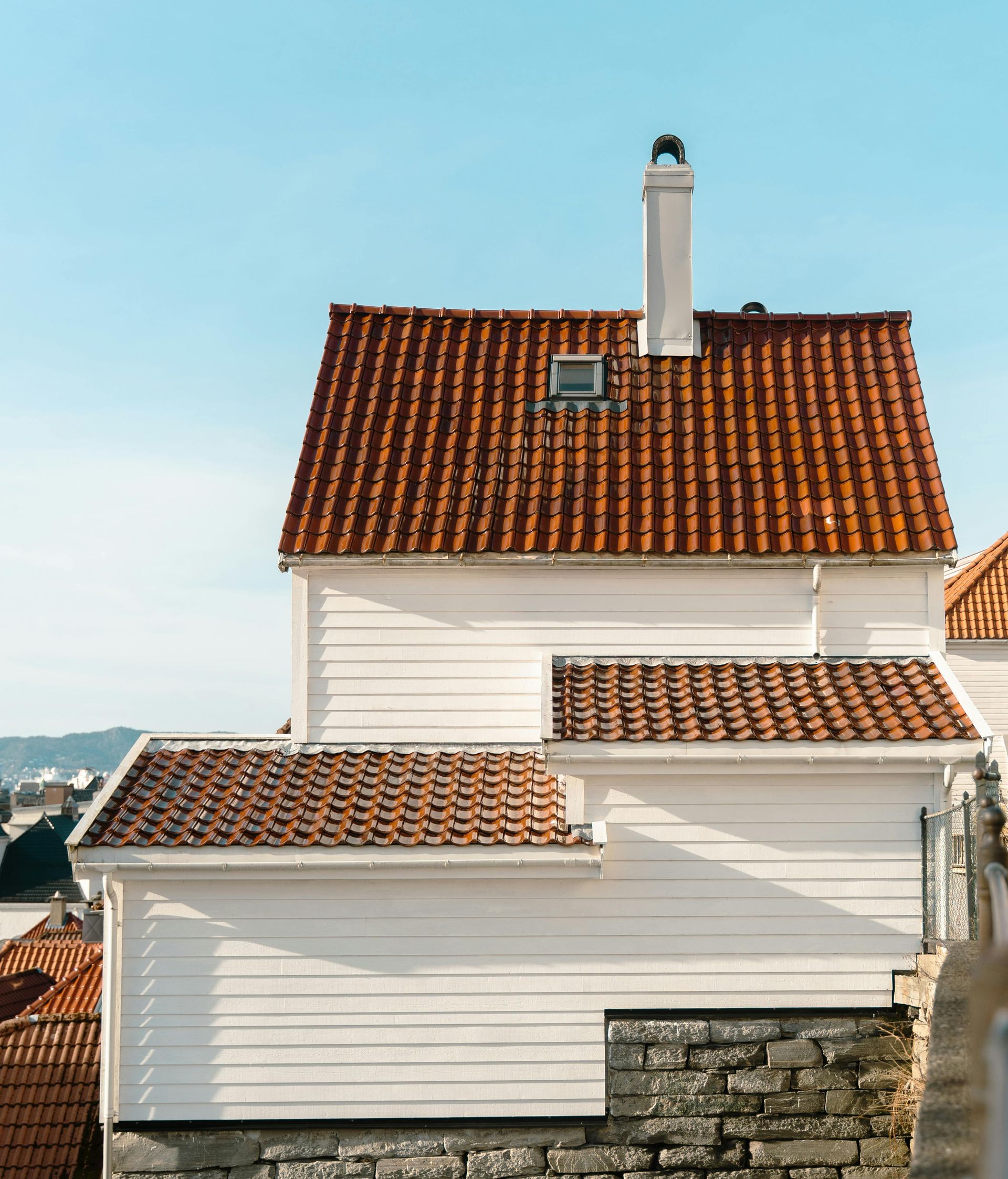 White house with red-tiled roof and chimney beside a stone wall under a blue sky
