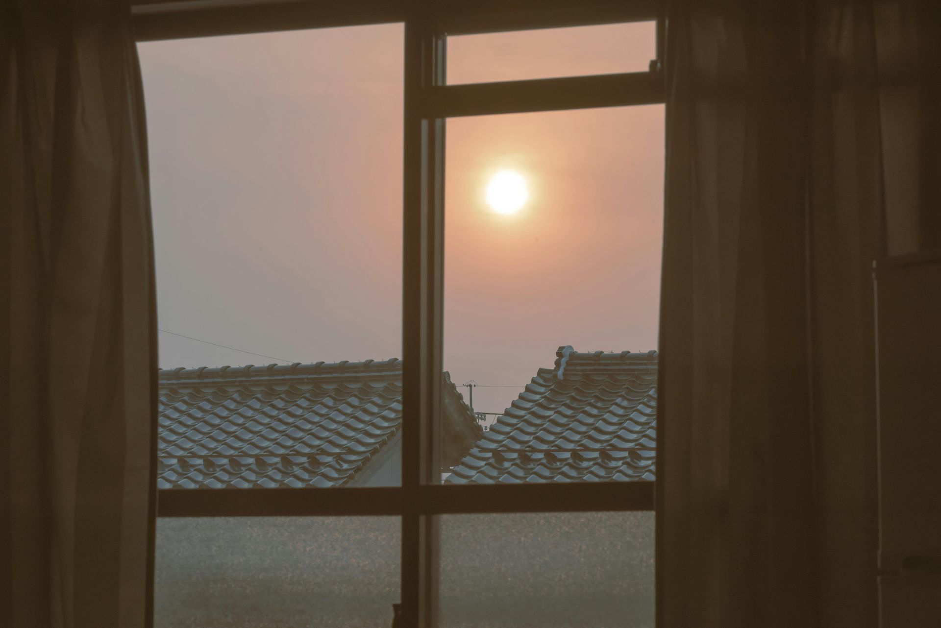 Sunset seen through a window over tiled rooftops, with curtains framing the view.