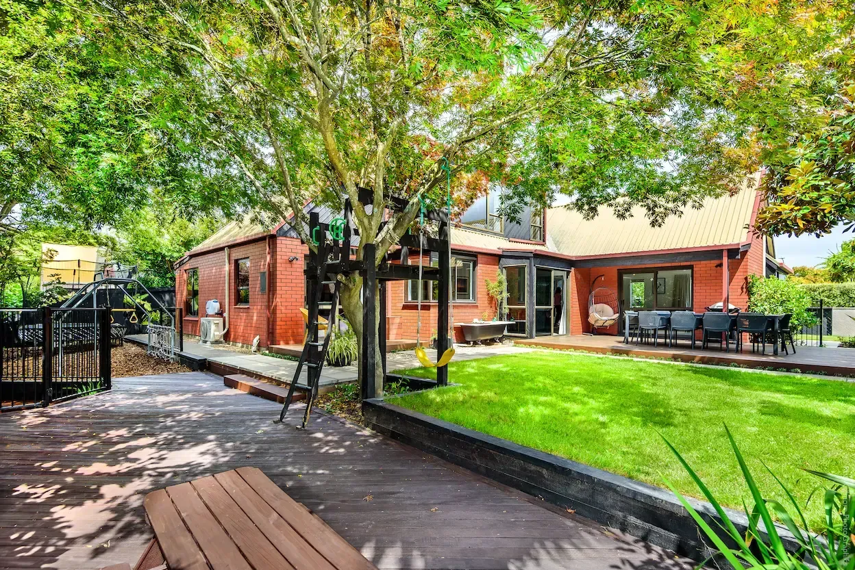 Red brick house with a wooden deck and green lawn shaded by a large tree.
