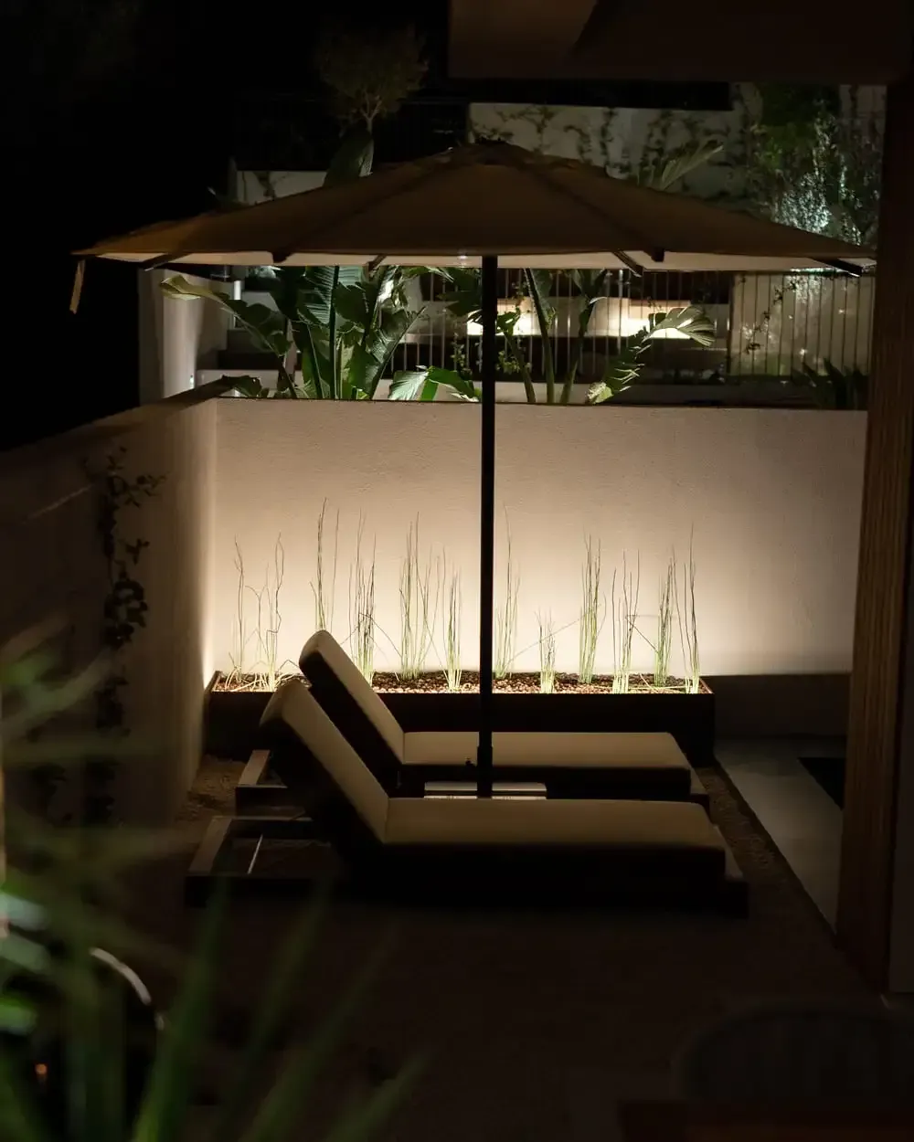 Gece aydınlatılan bir şemsiyenin altında iki şezlong. Arkada bitkilerle kaplı bir duvar var.