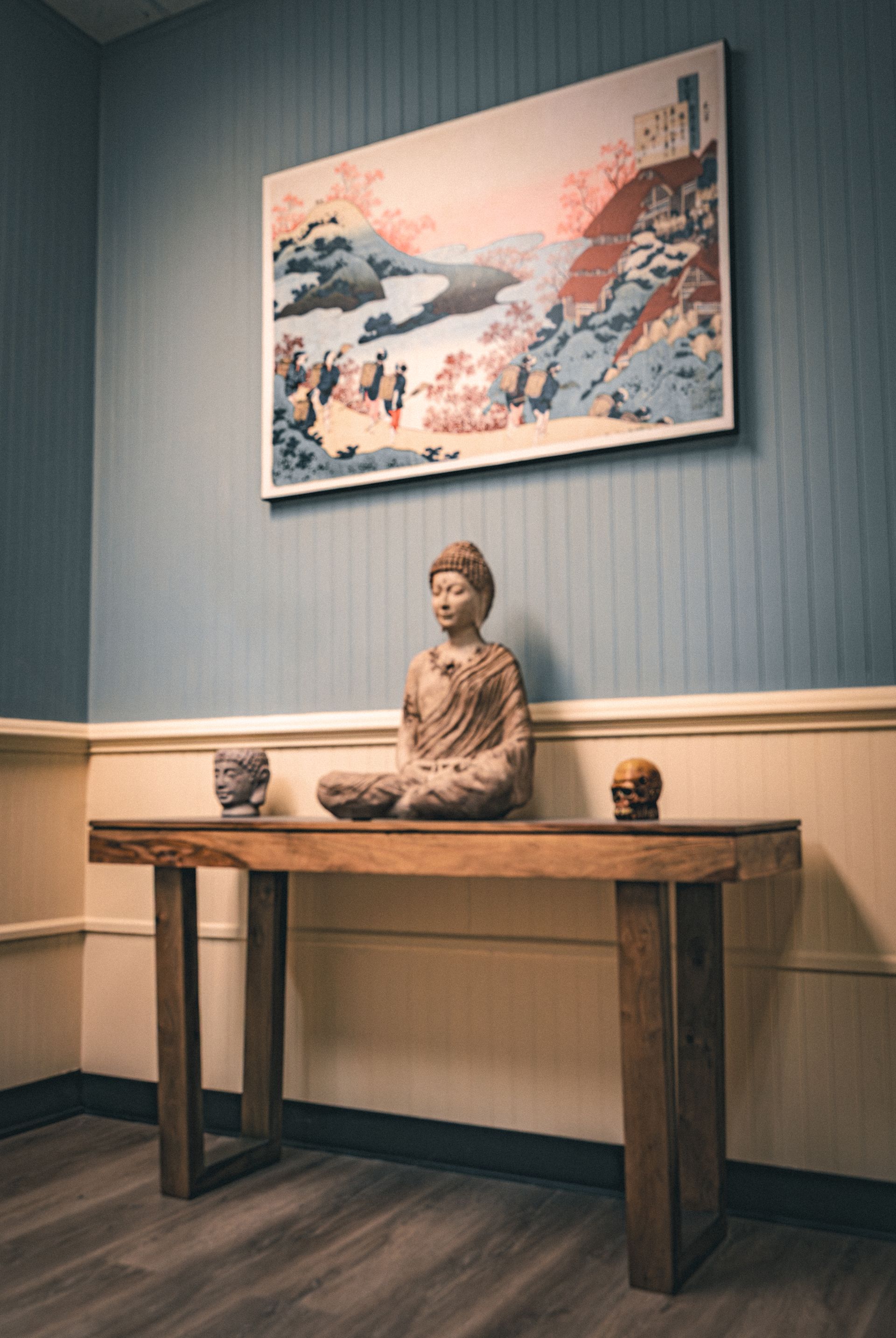 Buddha statue and decorative skulls on a wooden console table beneath a landscape painting in a blue and white room.