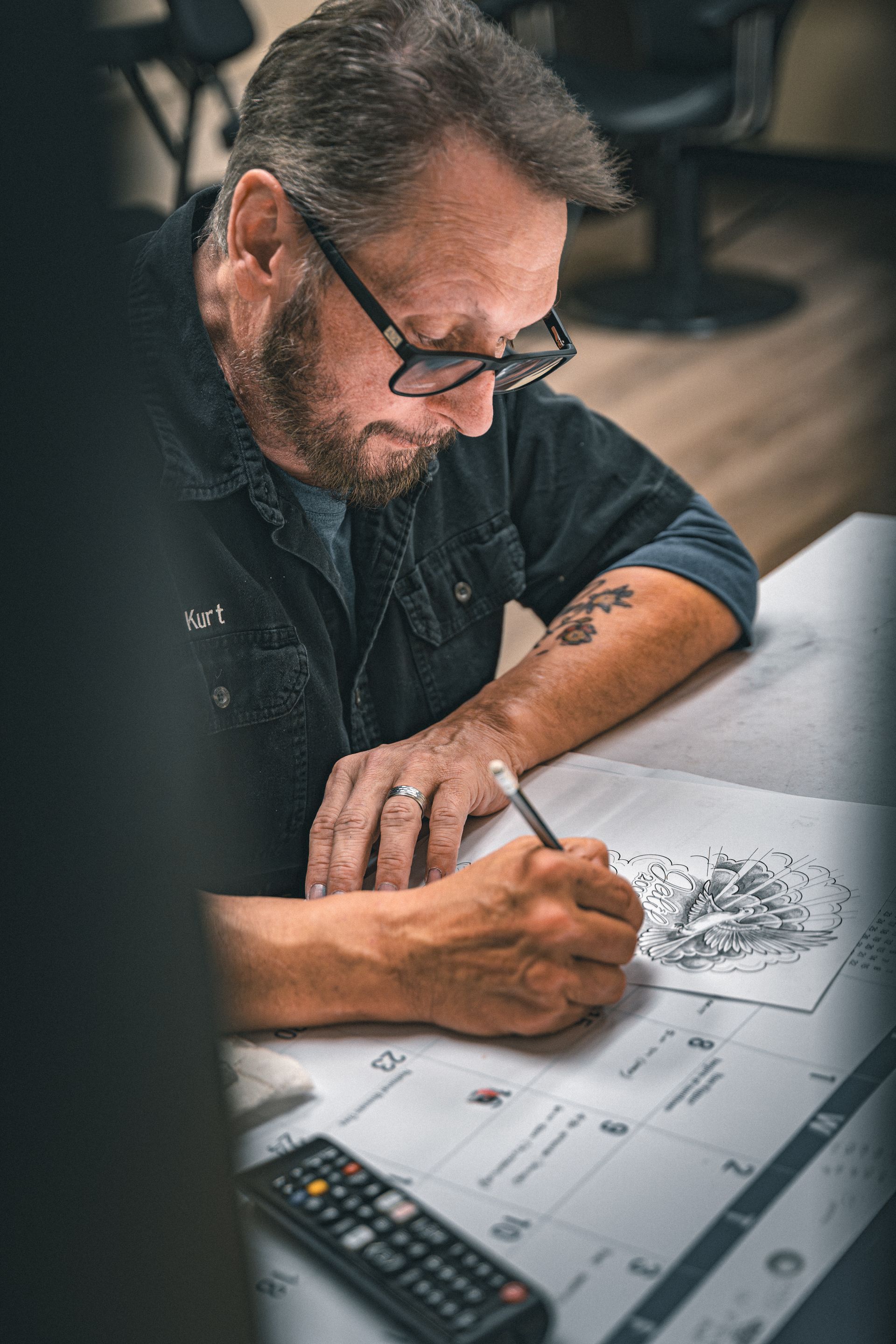 Man with glasses drawing, focused, using a pencil. Tattoo on his arm, working at a table, remote in the foreground.