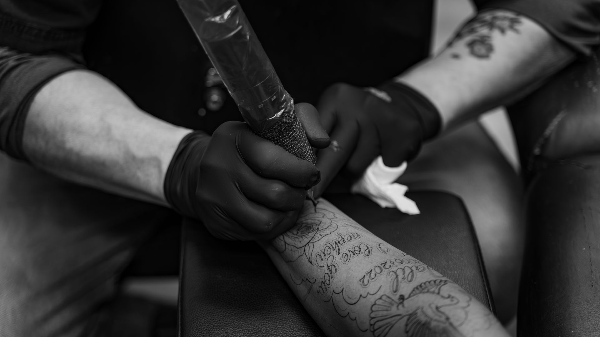 Tattoo artist with gloved hands working on a forearm tattoo; black and white.