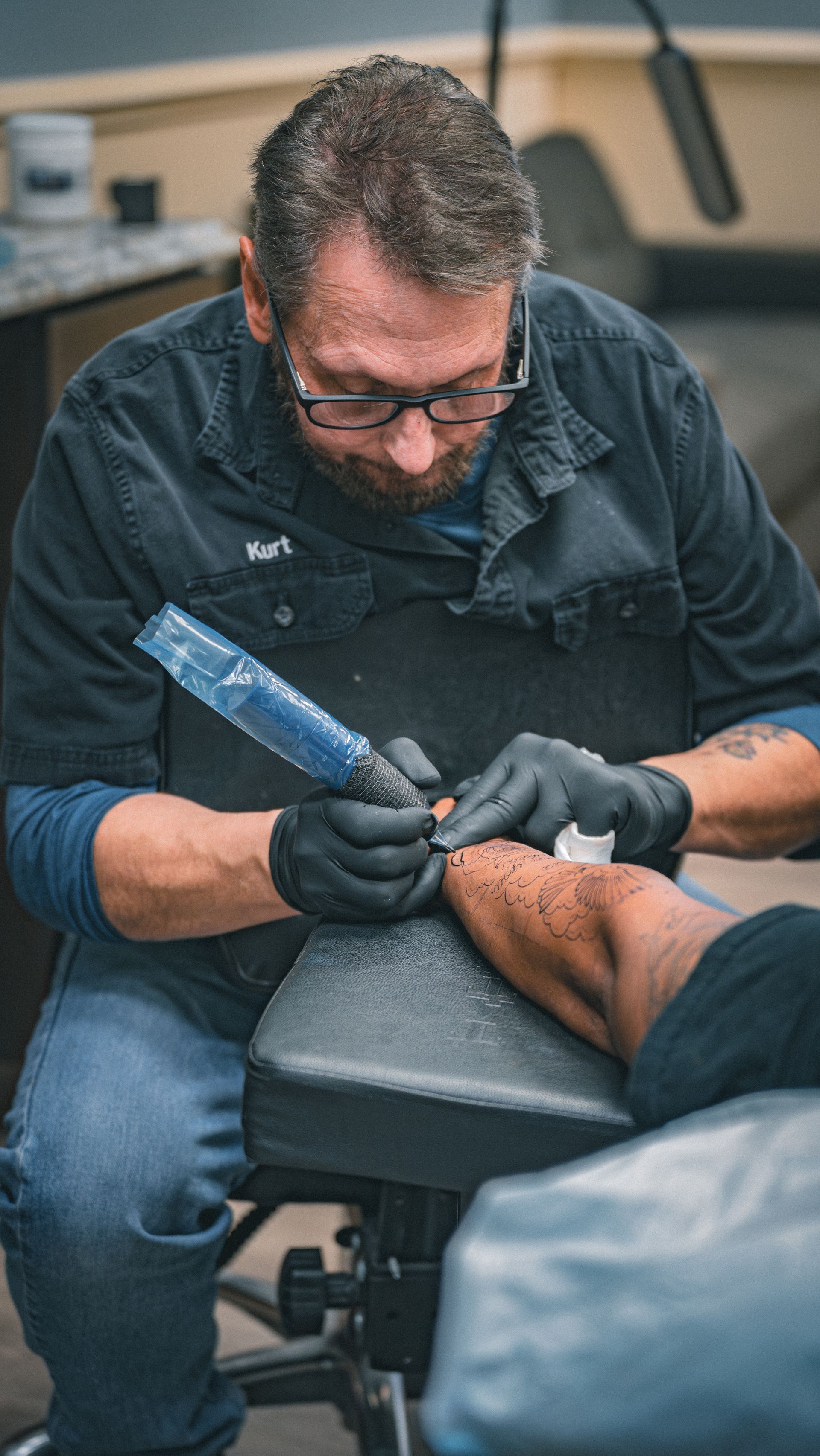 A person tattoos another person's arm. The tattoo artist wears glasses and gloves, working in a studio.