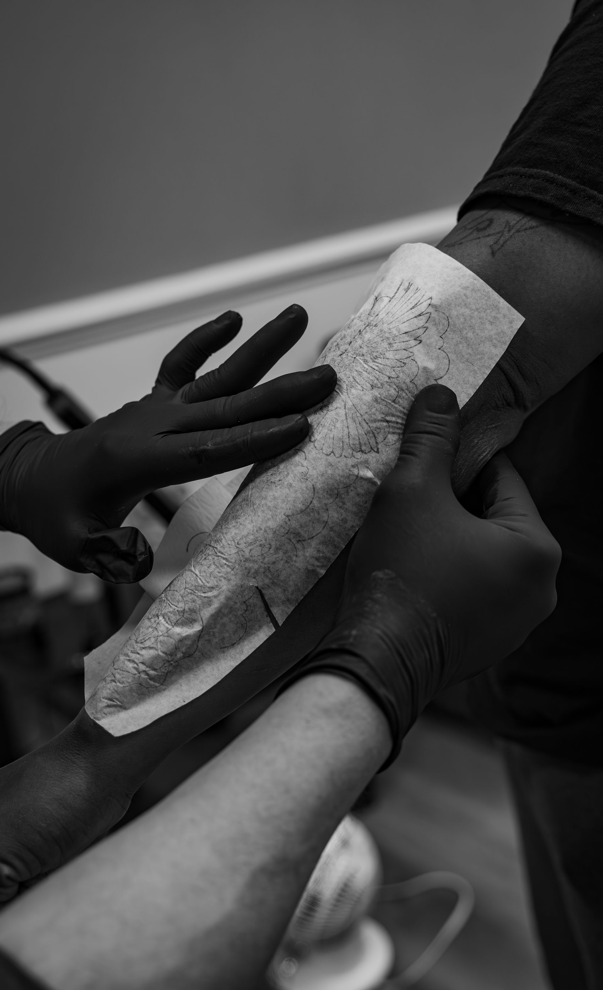 Tattoo artist's gloved hands applying a stencil to a client's arm. Black and white.