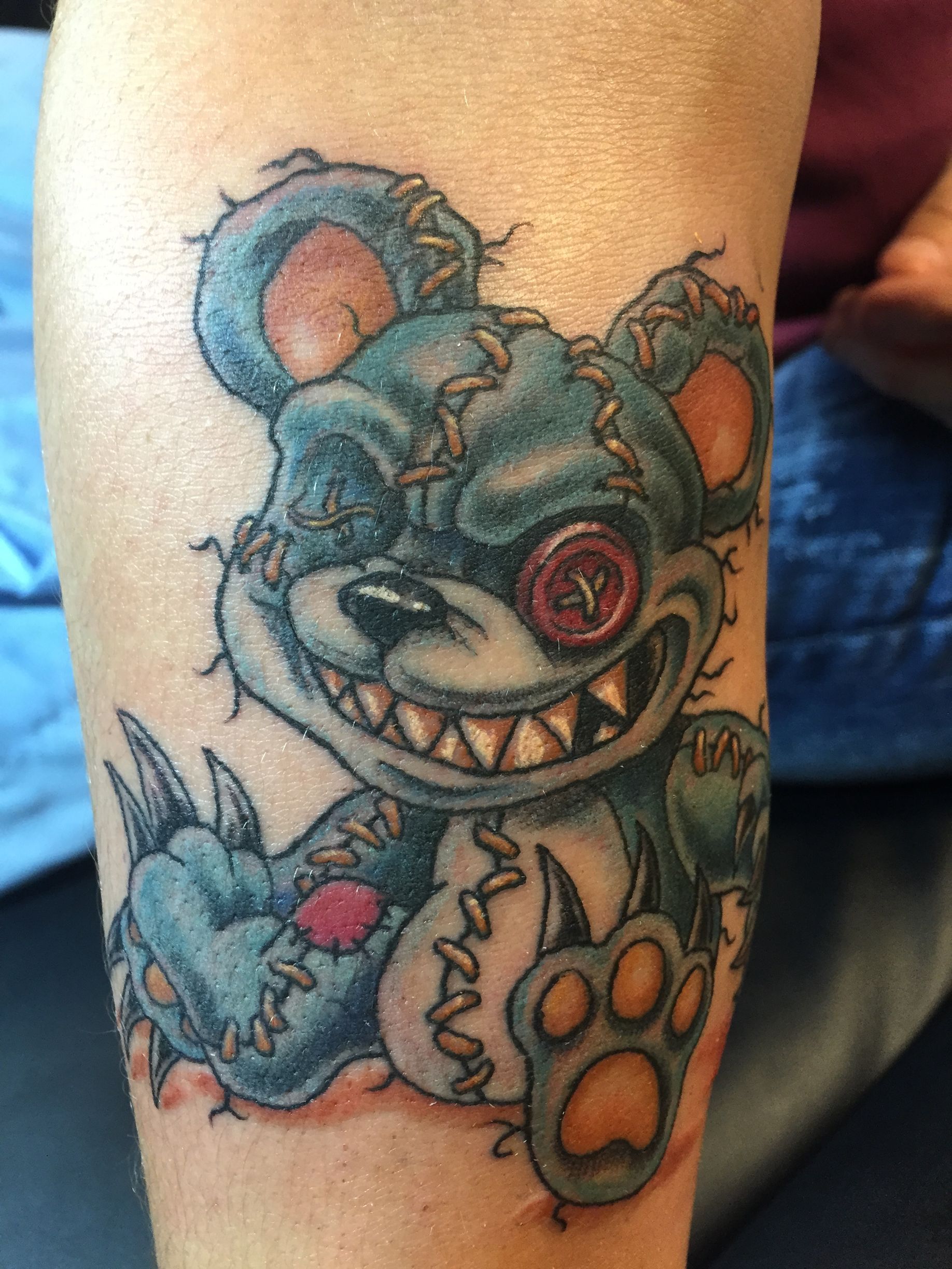 Tattoo of a stitched-up, evil-looking teddy bear with a button eye and sharp teeth, on a person's arm.
