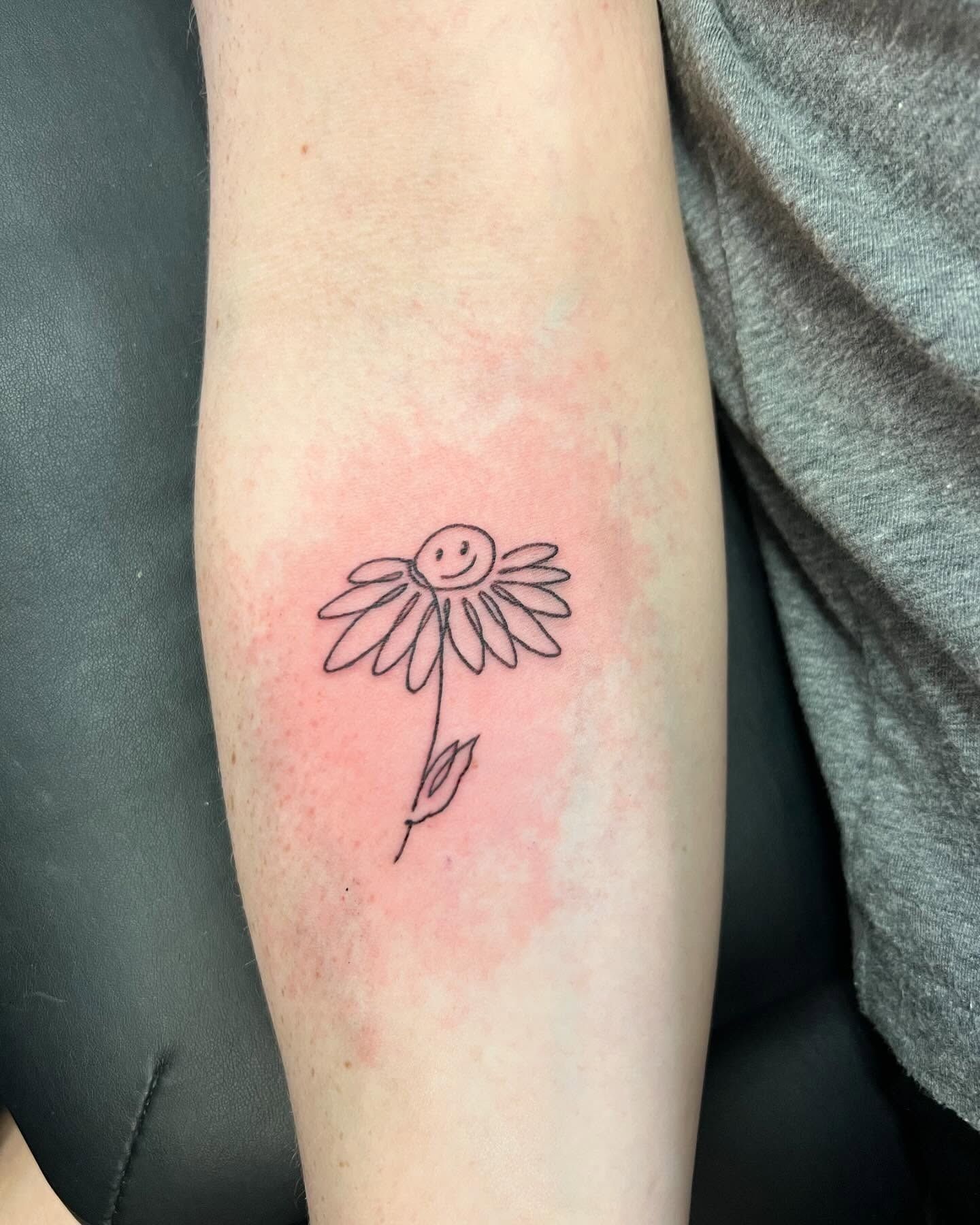 Line drawing tattoo of a smiling daisy on a forearm, surrounded by a pinkish-red rash.
