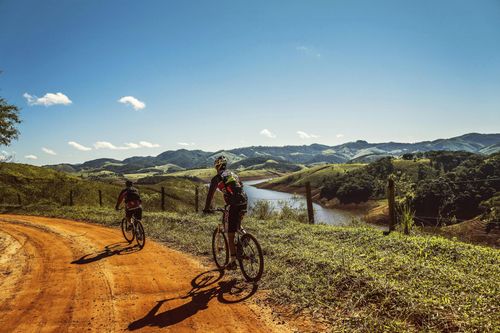 Two cyclists on a dusty trail overlooking green hills under a bright blue sky