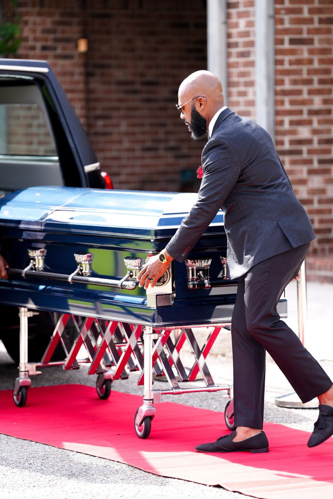 A person in a dark suit moves a blue casket on a rolling stand along a red carpet outdoors.