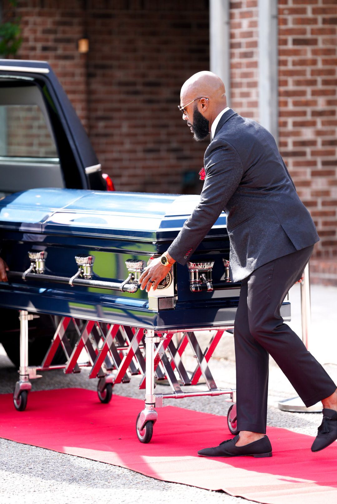 A person in a dark suit moves a glossy blue casket on a rolling metal stand along a red carpet.