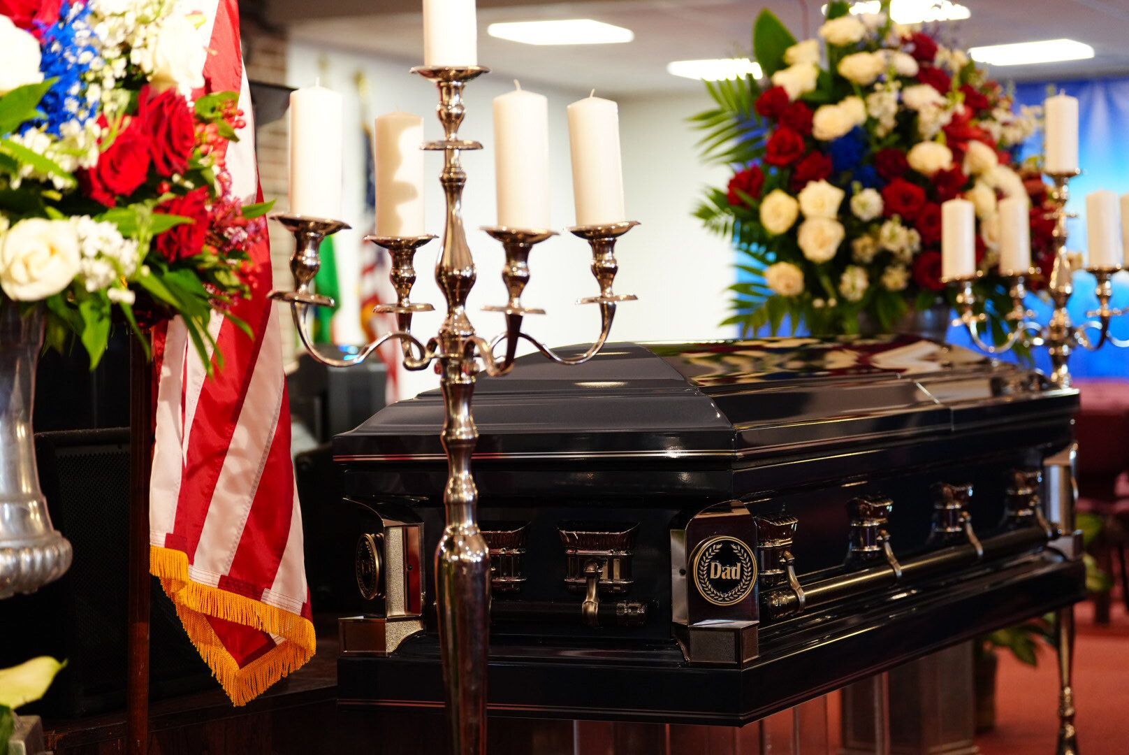 A dark casket sits in a room with flower arrangements, American flag decor, and silver candelabras.
