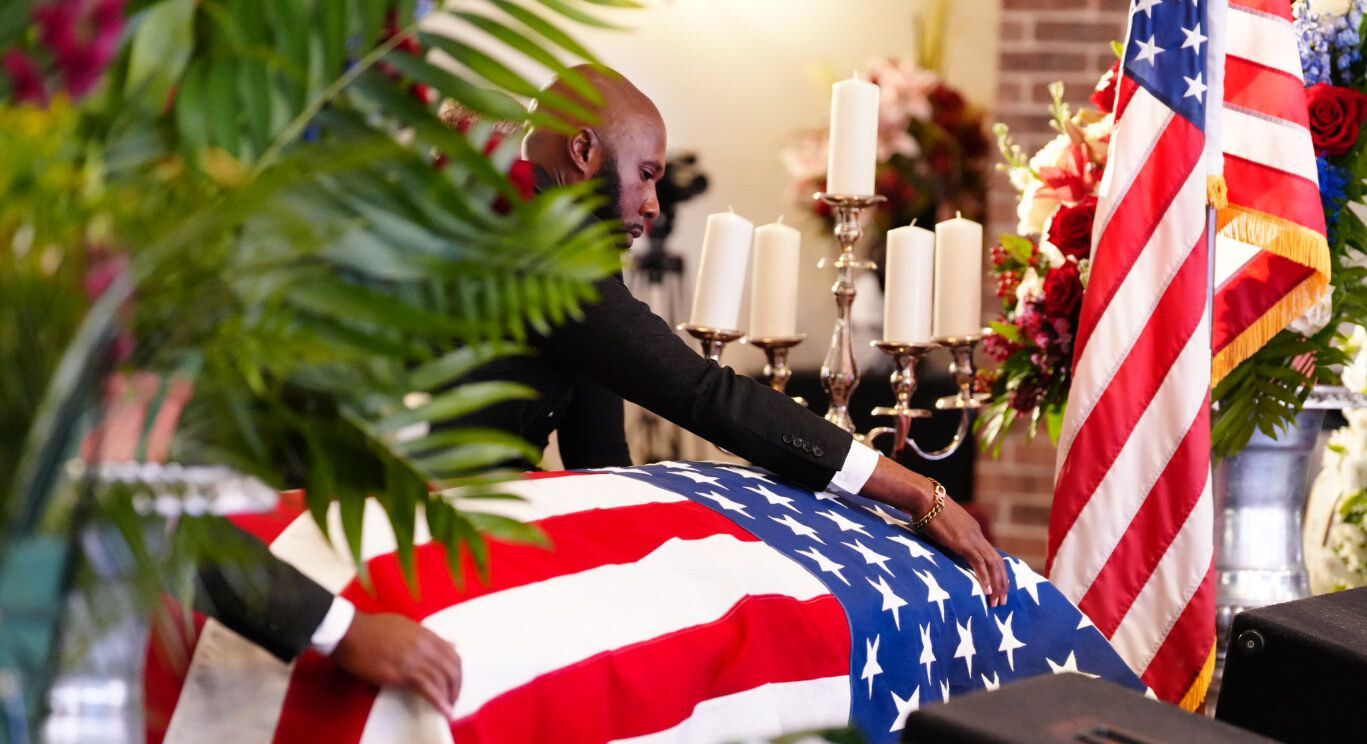 Hands rest on an American flag draped over a casket during a memorial service with lit candles and floral arrangements.