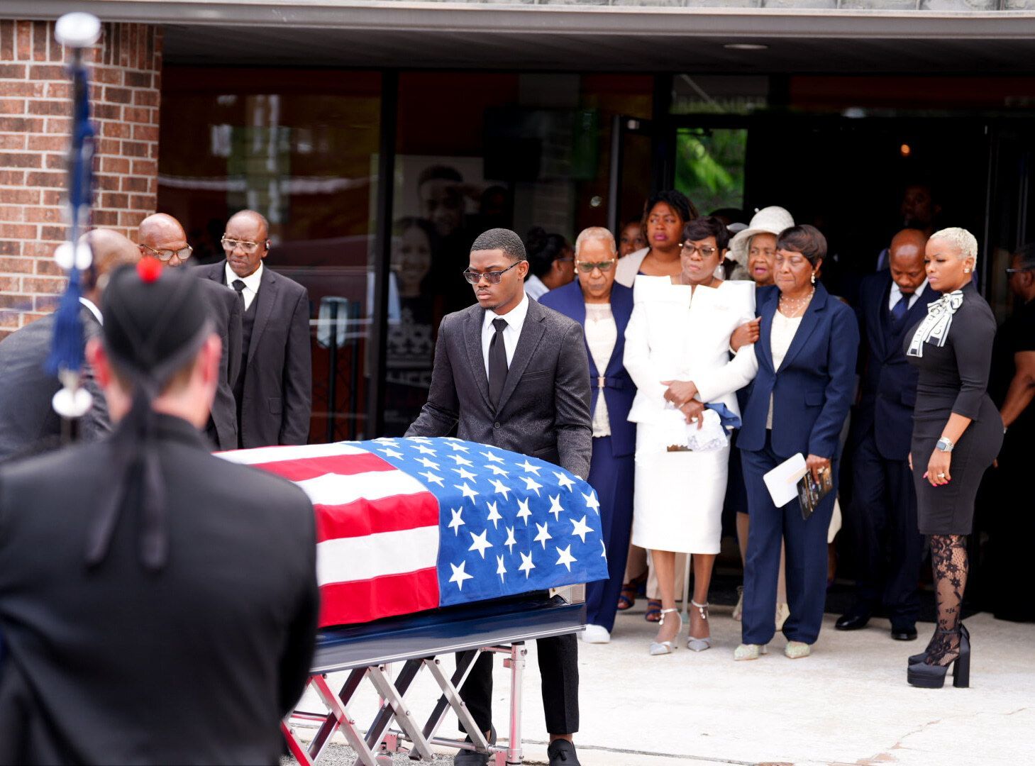Pallbearers carry a casket draped in an American flag out of a building, followed by a group of mourners in formal attire.