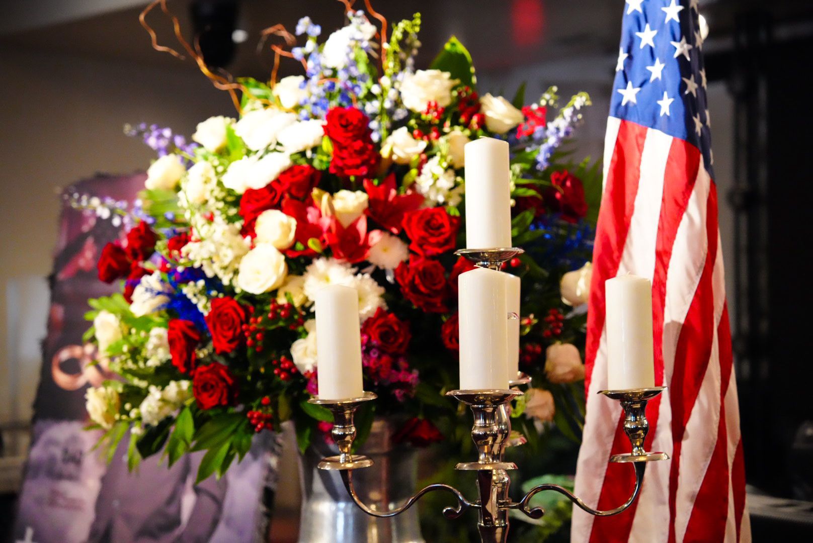 A silver candelabra with three unlit white candles sits before a patriotic floral arrangement and an American flag.