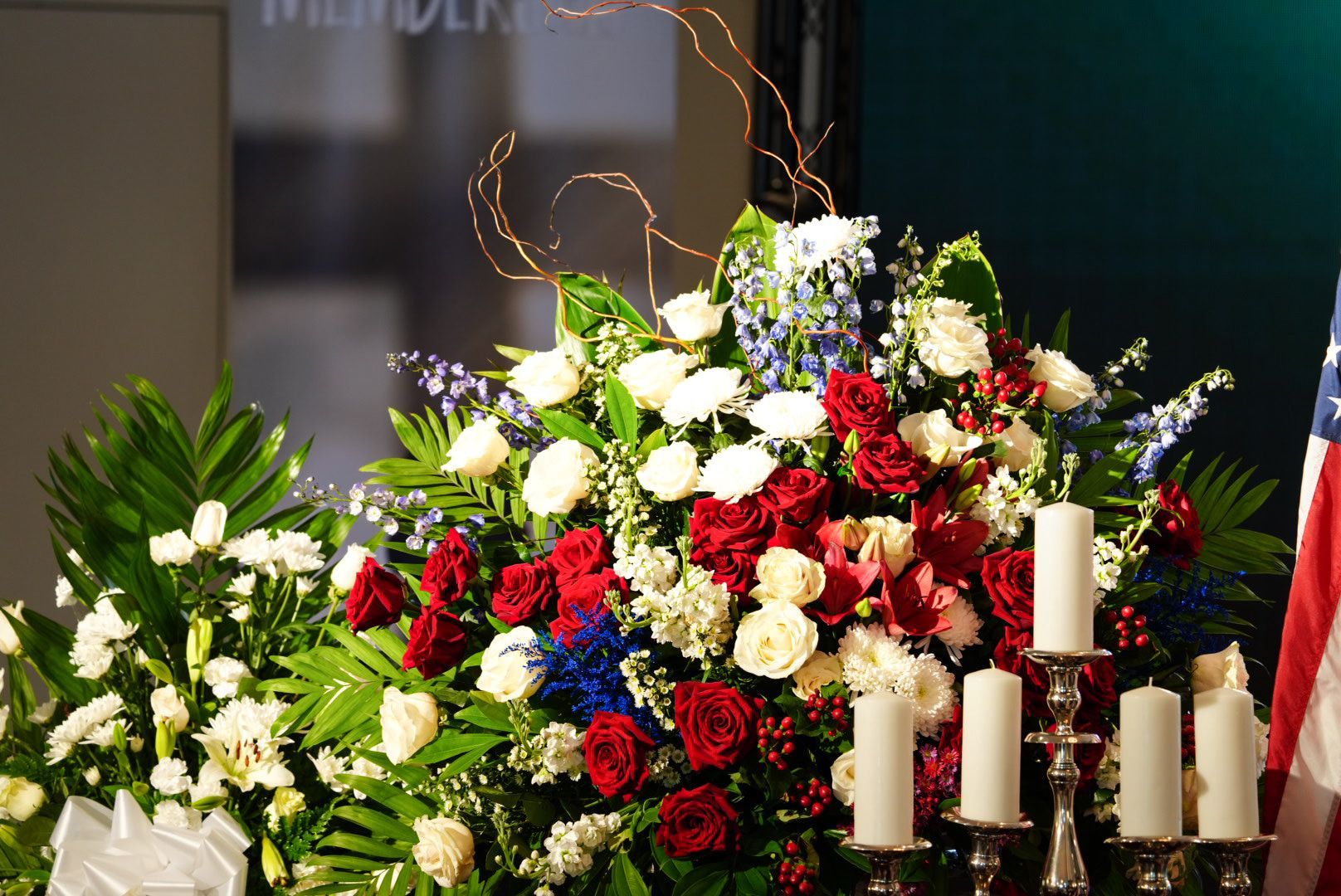 A large floral arrangement featuring red and white flowers and greenery next to several white candles and an American flag.