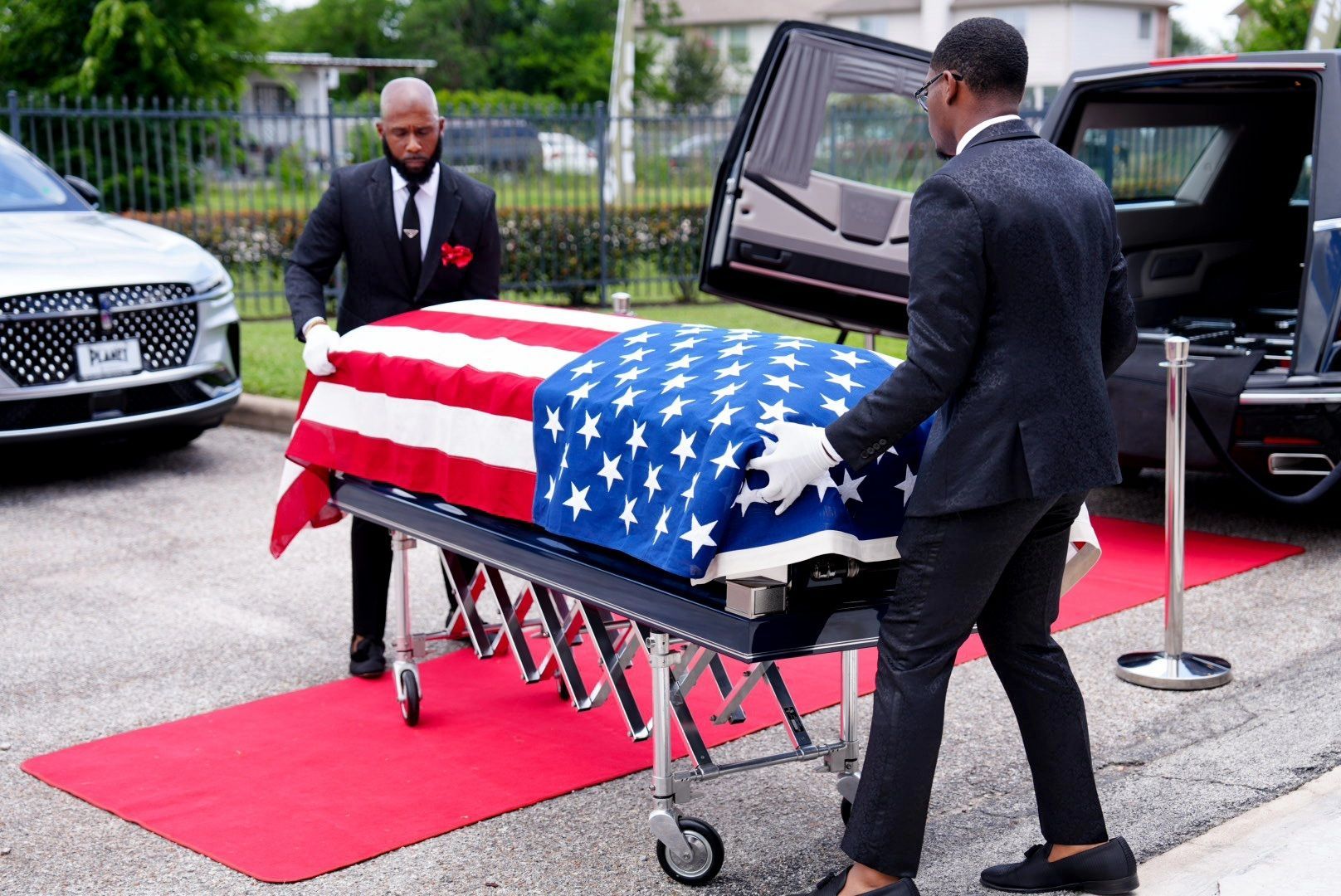 Two people in suits and white gloves move a casket draped in an American flag on a rolling cart toward a hearse.