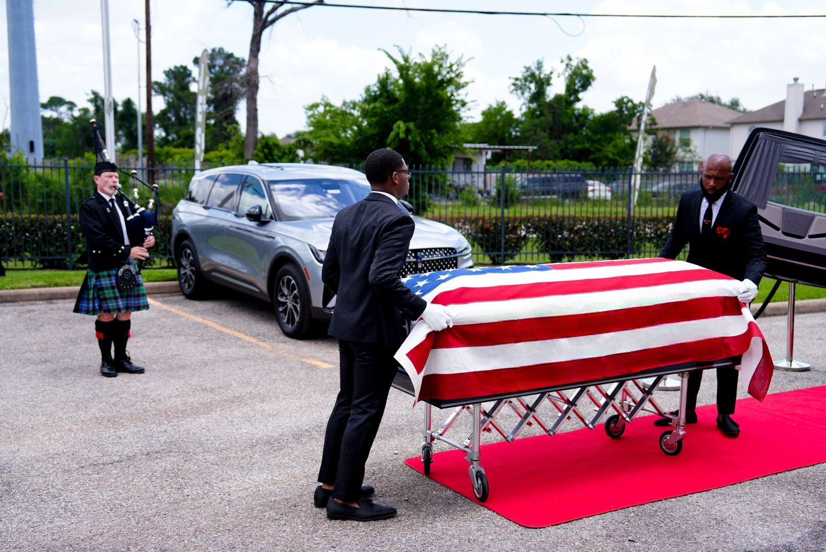 Two people in dark suits move a flag-draped casket on a red carpet while a bagpiper plays in an outdoor parking lot.