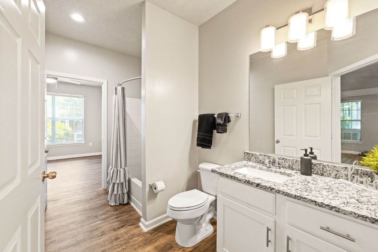 Bathroom in apartment featuring a granite countertop vanity, toilet, shower/tub with curtain, and doorway to adjoining room.