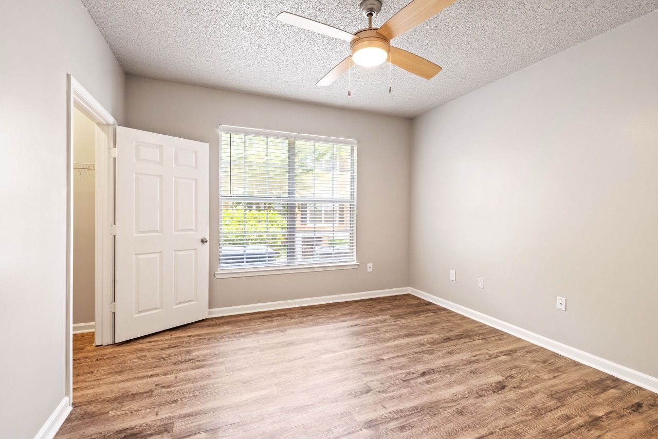 Empty bedroom in an apartment with a window, door, wood-flooring, and a ceiling fan.