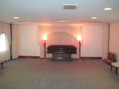 A funeral parlor with a closed casket centered; two candelabras flank it, against a draped backdrop; soft lighting.