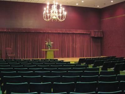 Rows of empty theater seats facing a stage with closed red curtains, a table, and a chandelier.