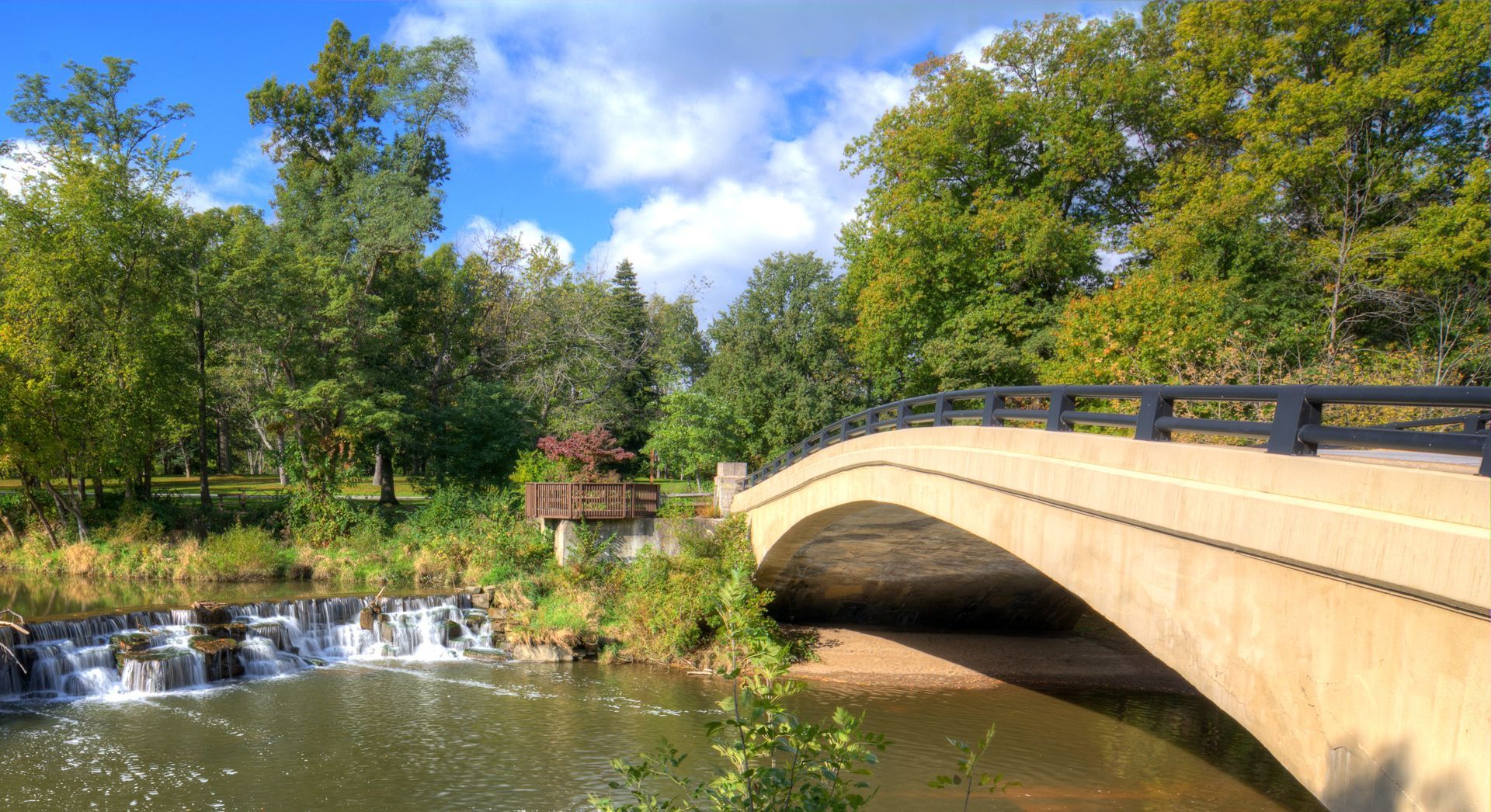 Bridge over a river with a small waterfall, trees, and blue sky.