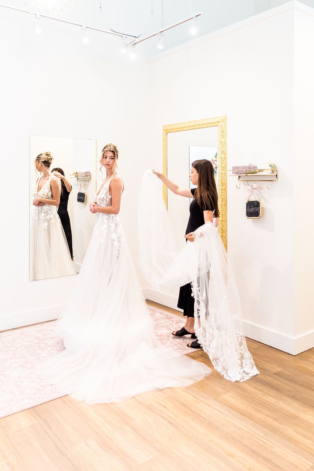 Bride in a wedding dress, looking at a mirror, with a person holding up a veil in a bridal shop.