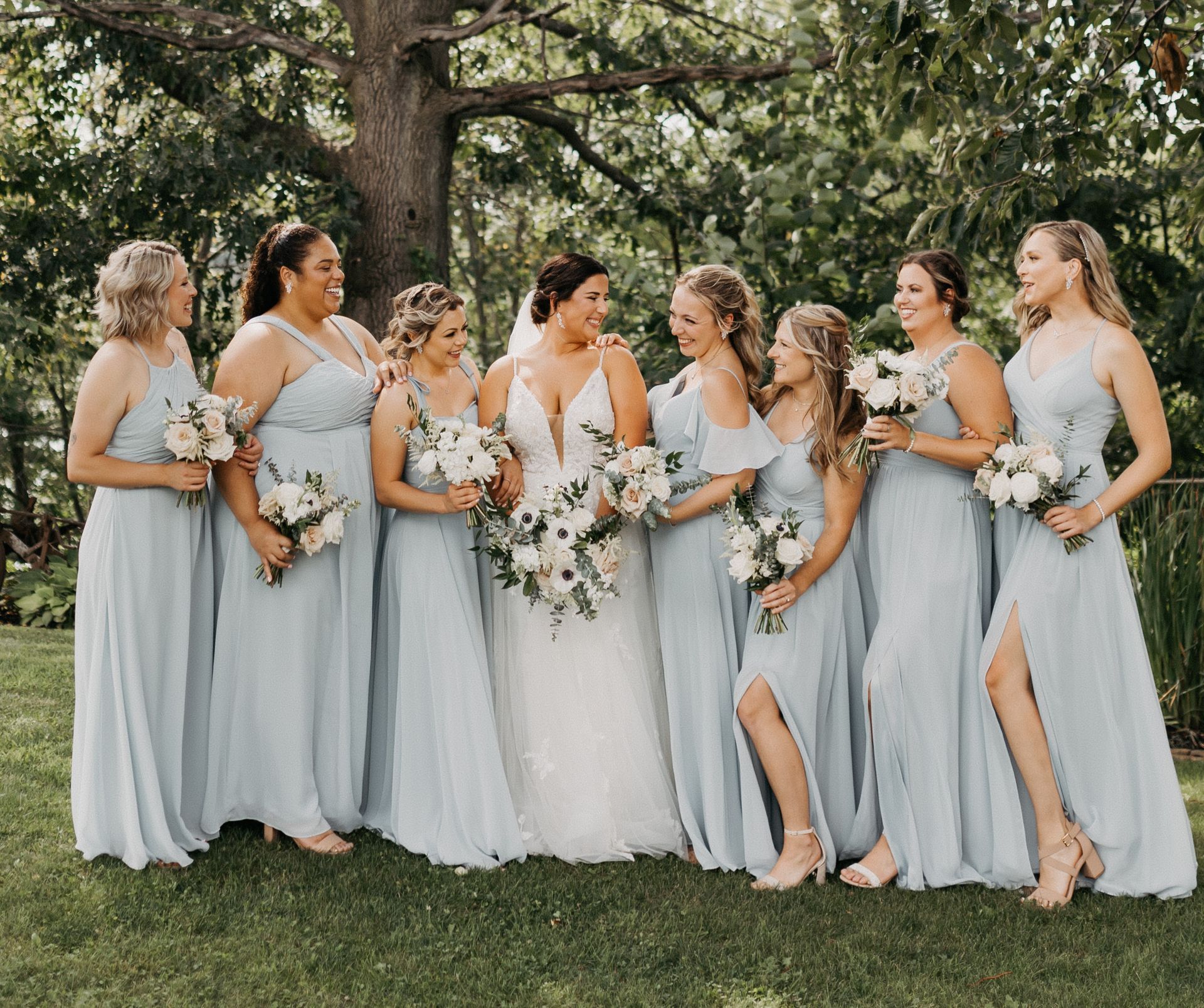 Bridesmaids in flowing dresses hold hands outdoors; a building is in the background. Dresses are shades of green and white.