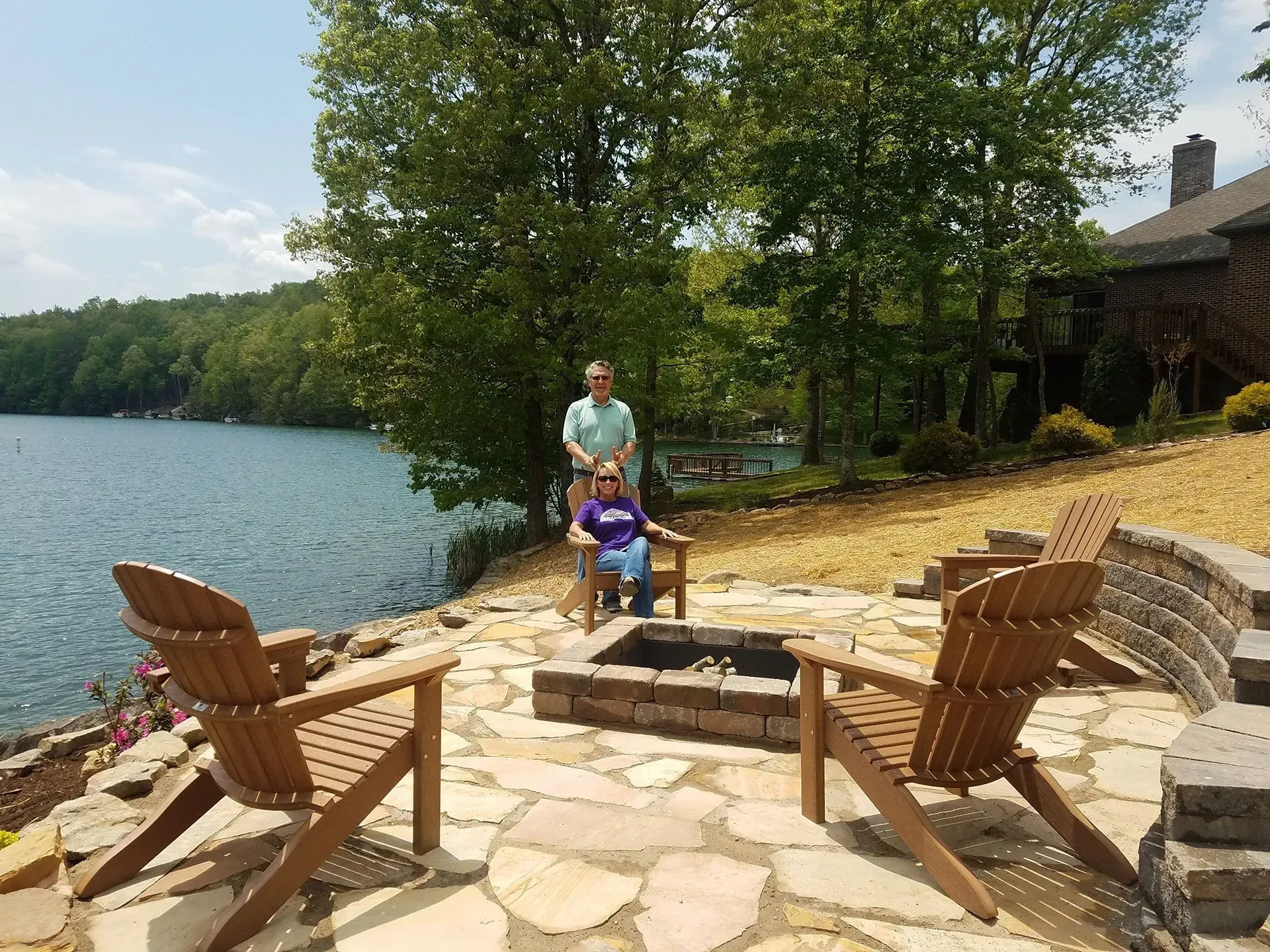A man and woman are sitting at a fire pit next to a lake.