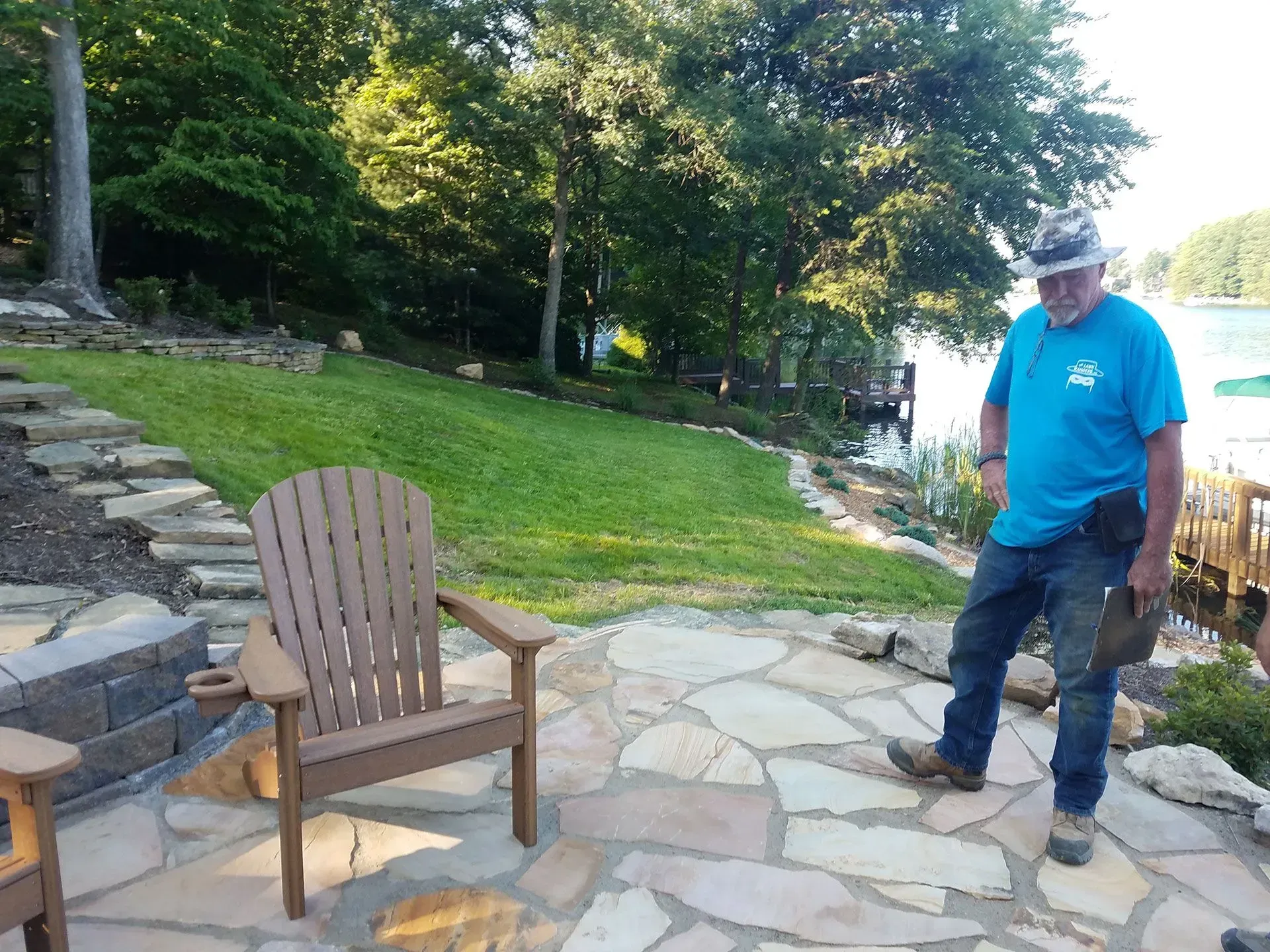 A man in a blue shirt is standing next to a wooden chair on a patio.