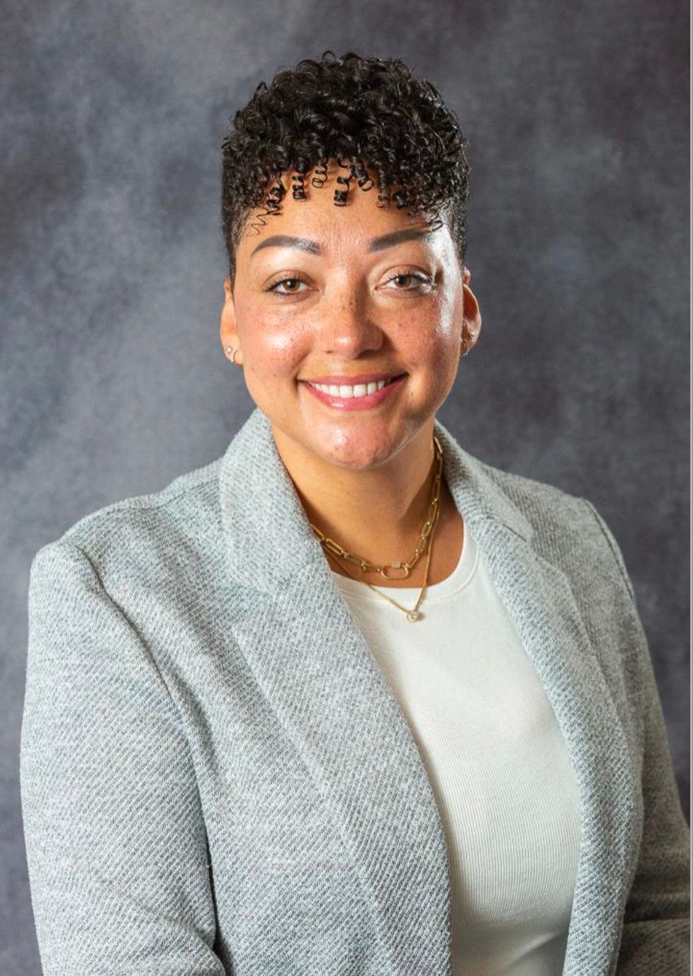 a woman in a grey jacket and white shirt is smiling for the camera .