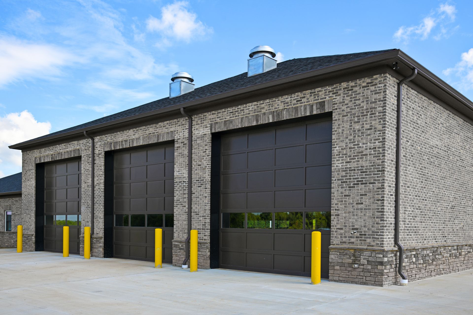 A brick building with three garage doors and yellow bollards. Chimneys on the roof.