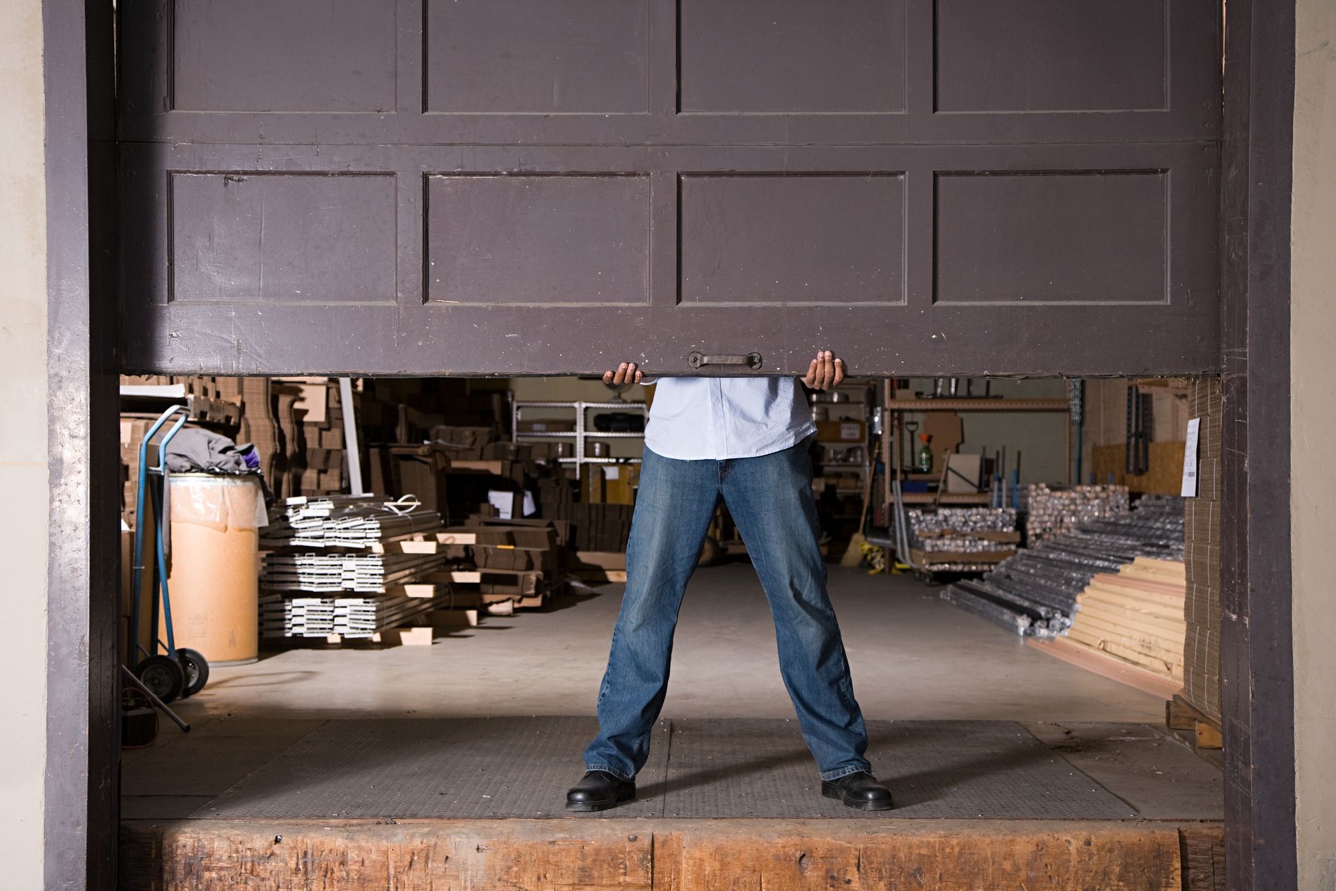 A person in a light blue shirt and jeans lifts a large, dark brown industrial garage door in a warehouse setting.