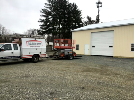 A man in blue overalls repairs a white garage door. A toolbox sits on the floor.