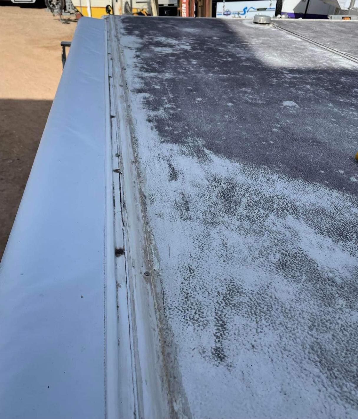 A close-up view of an RV roof, showing the edge and side with white trim. The roof surface is worn with debris.