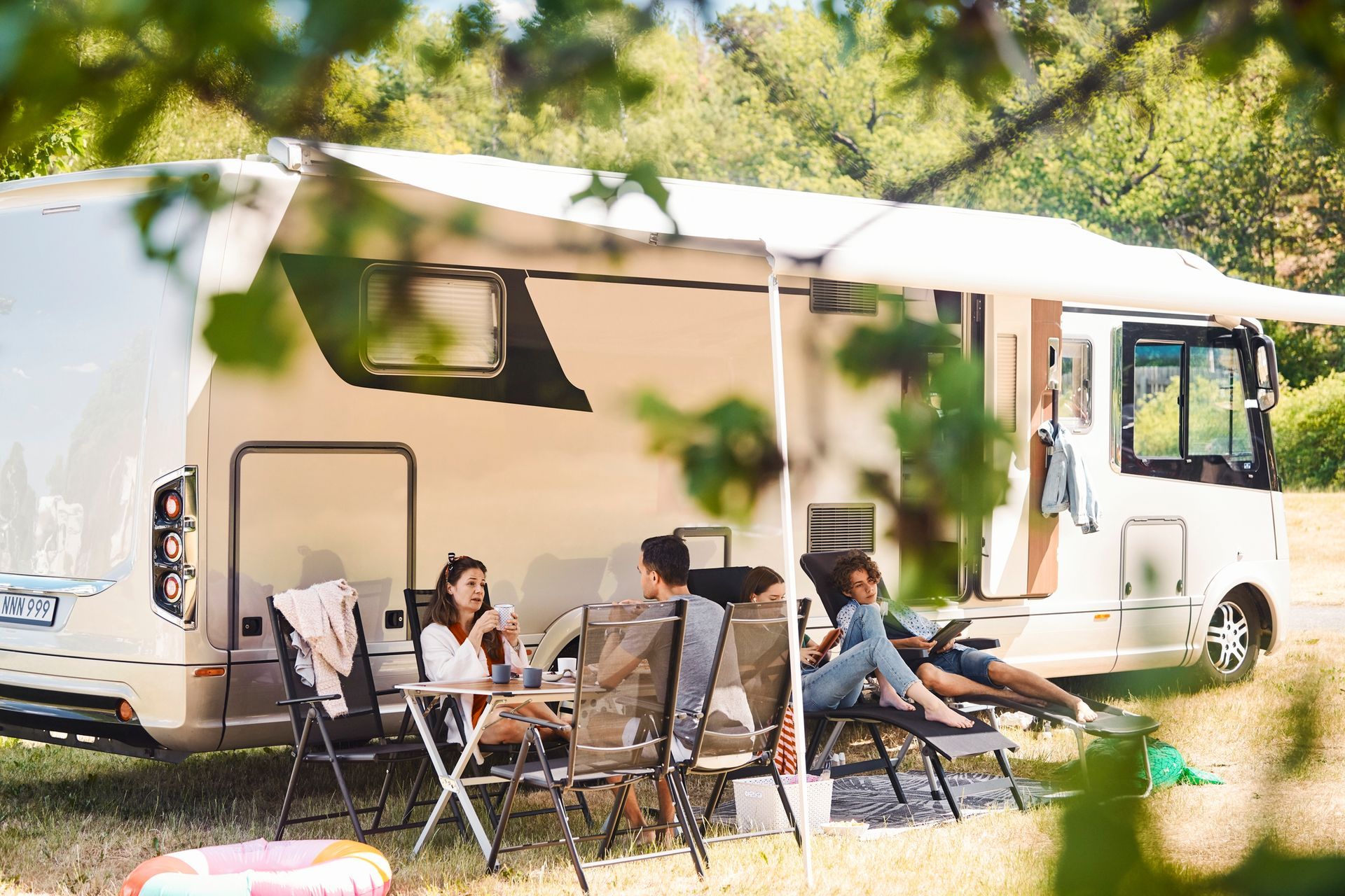 People relaxing outdoors beside a large RV with awning extended; sunny day.