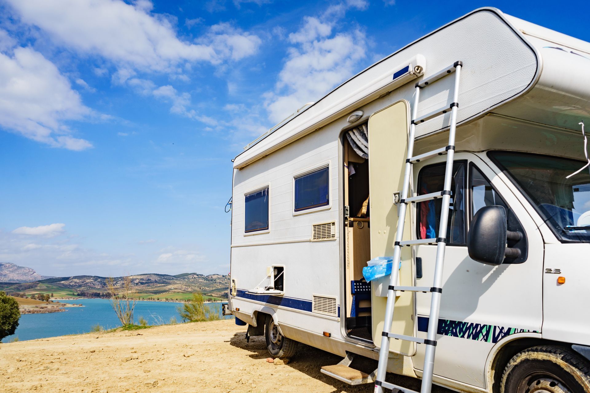 RV parked on a hillside overlooking a lake under a bright blue sky. Open door with ladder attached.