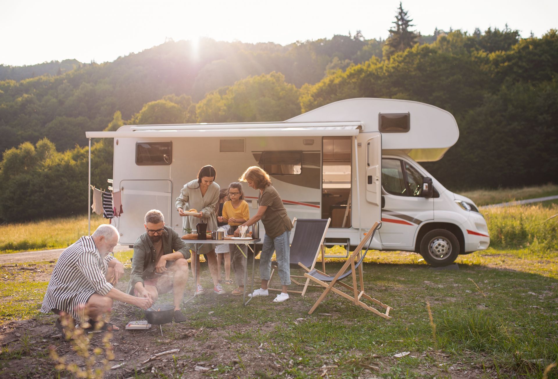 Family camping with a motorhome, enjoying food at a table near a forest, warm sunlight.