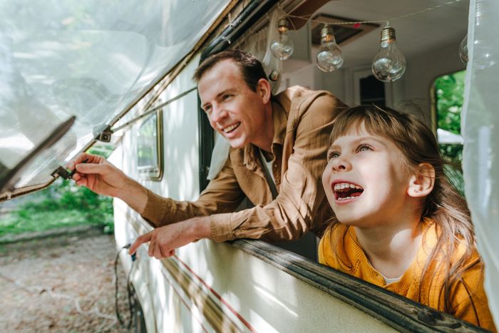 Man and girl look out RV window, smiling. Girl points. Green trees in background.