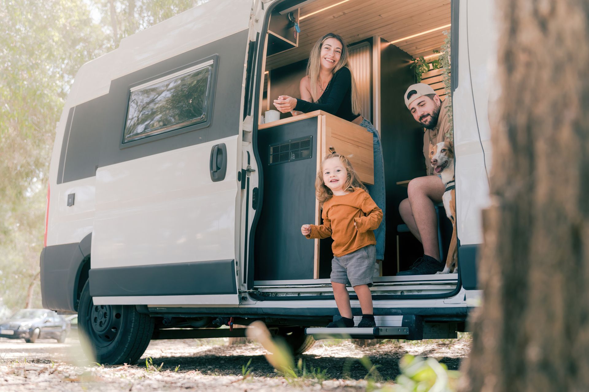 Family inside a camper van in a park. Smiling parents and a child. The van is white and gray.